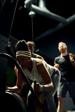 Diverse group training intense strength workout in gym with focused woman using cable machine. Her friends are cheering for her