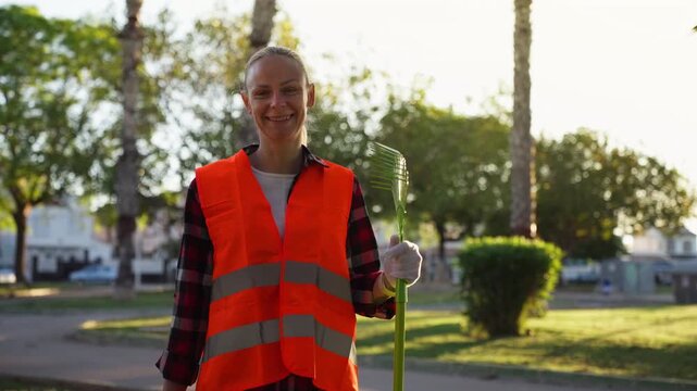 Positive female sanitation worker in bright orange safety vest smiling while holding a rake in sunny city park, representing public service, urban maintenance, and environmental care.