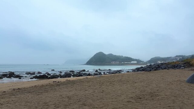 A gloomy view opens up over the sandy and rocky beach in Itoshima, Fukuoka. Dark rocks stretch out into the ocean under a thick blanket of grey clouds. 