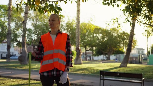Female environmental worker in safety vest cleaning public park with rake, promoting sustainability, environmental protection, and clean urban green spaces.