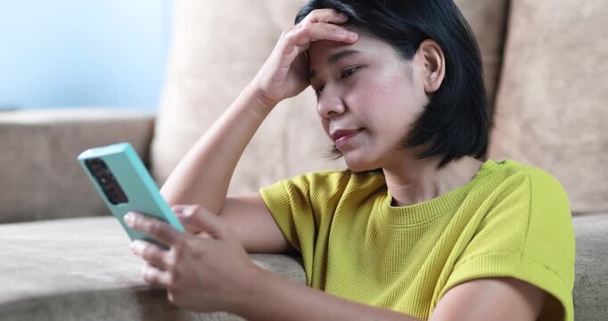 Stressed woman looking at smartphone while sitting at home. Concept of financial problems, bad news, debt, social media stress, anxiety, and emotional frustration in daily life