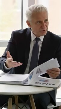 Vertical video: Picking up report, exec in suit, tie flipping chart pages by big window explaining