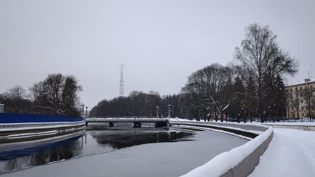 A partially ice-covered canal with concrete banks and a parapet runs between the trees. The ground is covered with snow, a well-trodden path. Nearby there are buildings and a bridge over the water