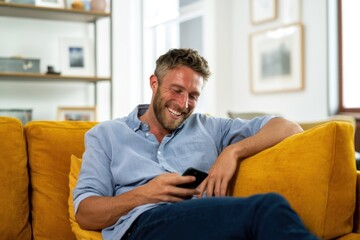 Fototapeta premium The man on a yellow sofa enjoying a laugh while using his smartphone at home