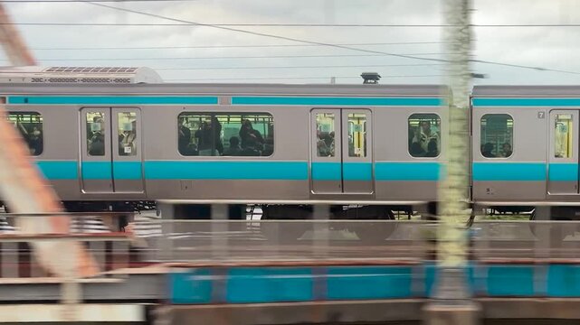 A passenger train passes over a steel truss bridge in Japan. The commuter train moves steadily on a multi-track railway line. High angle view shows suburban infrastructure with green riverbanks.