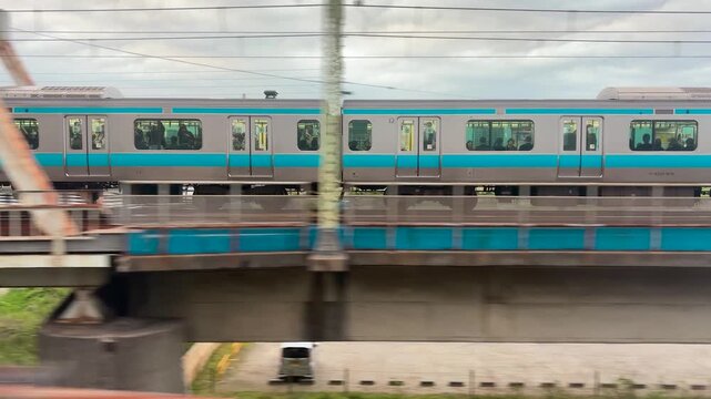 A passenger train passes over a steel truss bridge in Japan. The commuter train moves steadily on a multi-track railway line. High angle view shows suburban infrastructure with green riverbanks.