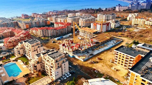 Aerial view of resort residential building construction site near sea. Drone shot of hotel or apartment complex under construction with tower crane near coastline in sunny weather.