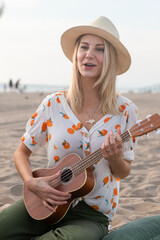Fototapeta premium Woman in a hat playing ukulele on the beach, smiling and relaxed.