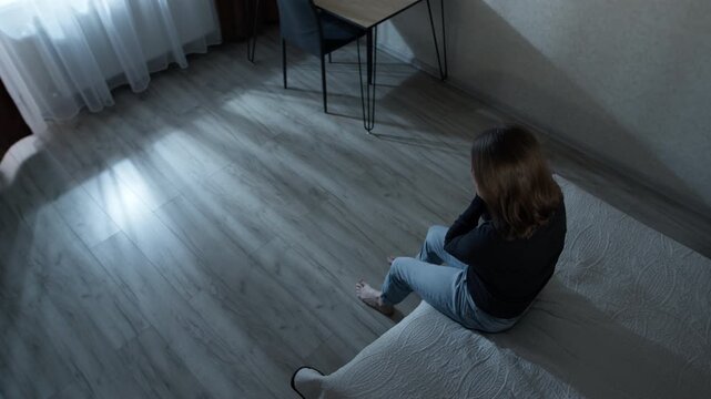 Lonely woman sitting on edge of bed during night. Depressed female suffering from insomnia, anxiety or psychological trauma inside dark bedroom.