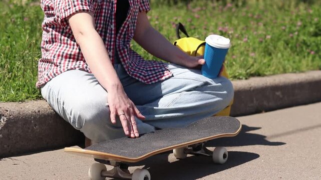 Unrecognizable person in comfortable summer clothes with skateboard. Young girl resting on the side of the road with skateboard. Concept of youth culture, sports and recreation