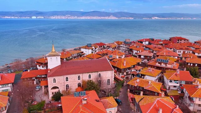 High angle view of Nessebar orthodox church and terracotta roofs. Aerial view of a stone church with white tower and golden spire among traditional red tiled houses by the sea.