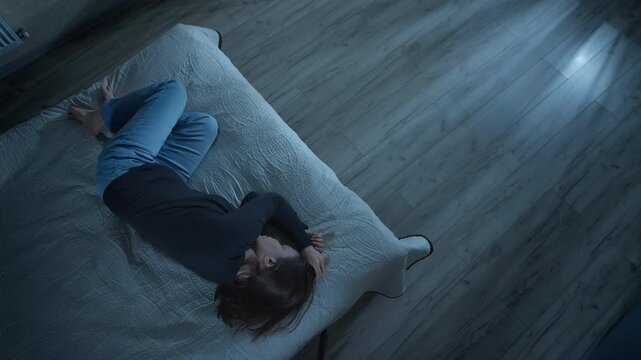 Top view of lonely woman lying in fetal position on bed during night. Depressed female suffering from insomnia or psychological trauma inside dark bedroom.