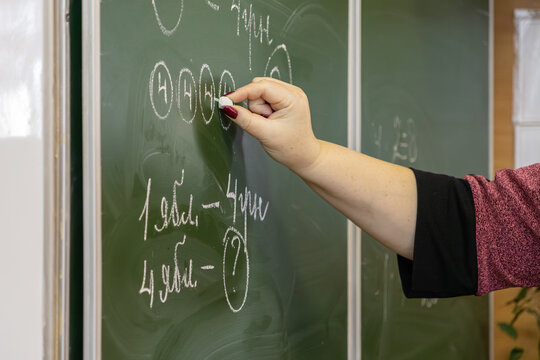 A teacher writes a math problem with chalk on a green chalkboard to explain elementary school arithmetic to students during a primary school lesson, fostering academic development now