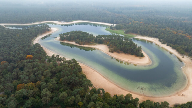 Aerial view of Henschotermeer lake with its central island, sandy beaches, and surrounding dense pine forest under a misty sky in Woudenberg, Utrecht, Netherlands.