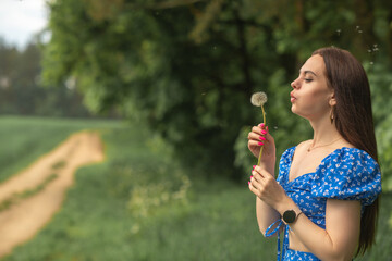 Fototapeta premium Young woman with long brown hair in blue floral dress blowing dandelion seeds in a green field, soft focus background with pathway and trees visible