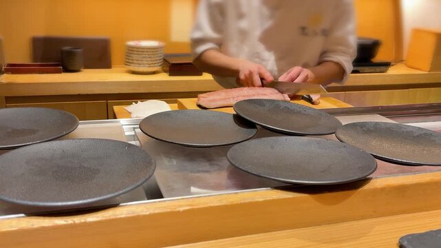 Professional Japanese sushi master slicing fresh raw tuna fish fillet on wooden cutting board in Tokyo. Experienced chef using authentic single bevel yanagiba knife to make cuts for premium sashimi.
