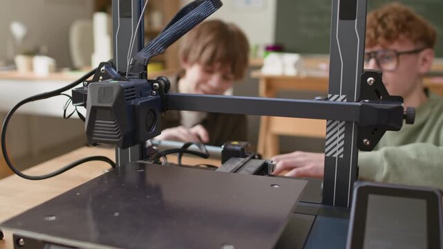 Close up shot of 3D printers extruder and guide rails, while two students setting up equipment and discussing printing process during school class