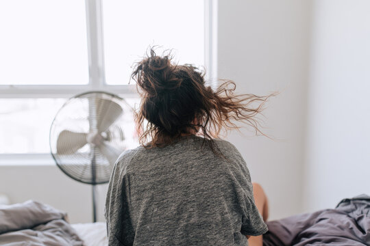 Bedroom electric fan cooling woman messy hair back view bright minimalist interior window light overexposed air flow comfort