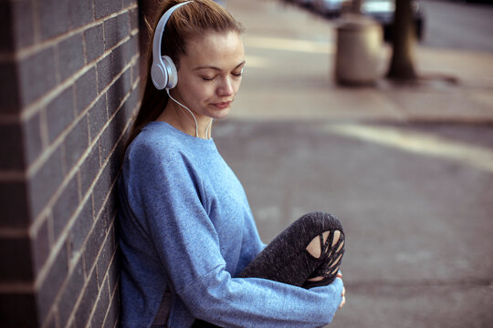 Young woman listening to music on headphones while sitting against a brick wall outdoors