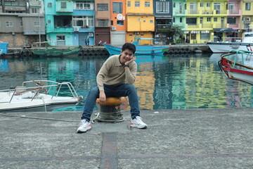 A man wearing a brown sweater and jeans stands posing by the pier with a view of colorful buildings in the background. © Peeb Kanom