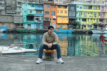 A man wearing a brown sweater and jeans stands posing by the pier with a view of colorful buildings in the background. © Peeb Kanom