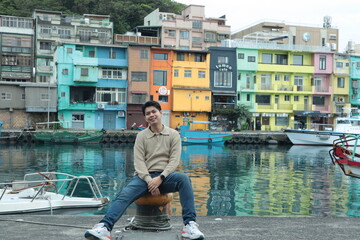 A man wearing a brown sweater and jeans stands posing by the pier with a view of colorful buildings in the background. © Peeb Kanom