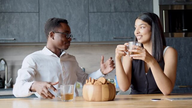 A black man and an Indian woman engage in light banter while enjoying tea and cookies during their break at the office. Their smiles show a budding connection as they interact