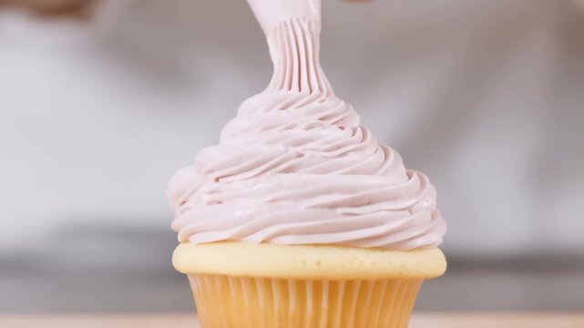Slow motion close up of hand piping pink frosting onto vanilla cupcake on kitchen countertop