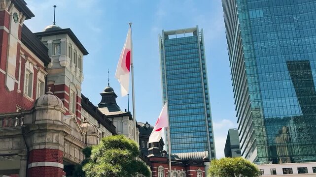 Japan flag waving near the historic Tokyo Station building in Marunouchi district. Red brick facade of the landmark railway station stands next to modern glass skyscrapers.