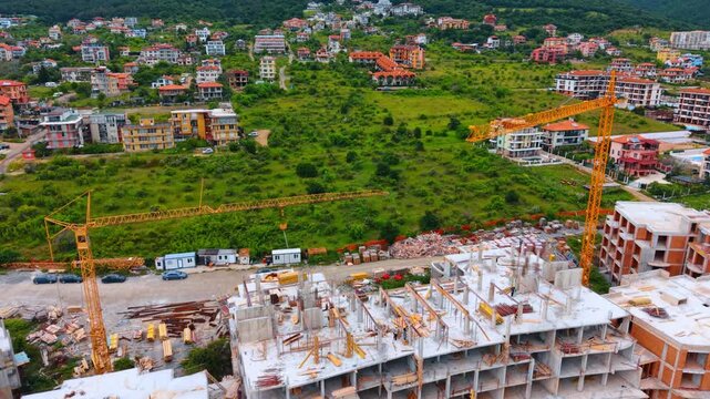 Construction site of multistory residential building with yellow cranes. Aerial shot of a concrete frame building under construction with two yellow tower cranes against a green hill background.