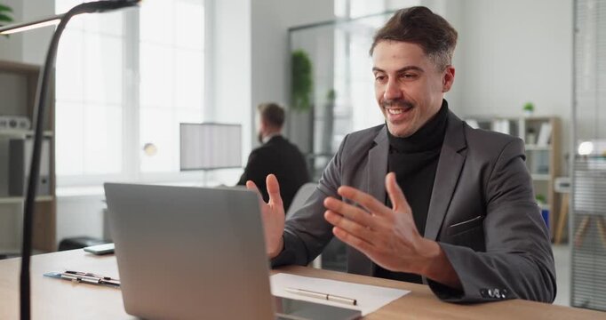 Businessman on video call in office. He smiles, waving during an online meeting with a client via laptop, discussing finance in a bright remote workspace. Concept of remote business meeting.