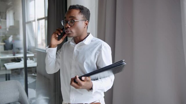 A man in an office talks on the phone. He is upset and raises his voice. He holds a black folder and seems frustrated because others are not following instructions on time