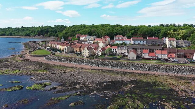 Reverse drone shot of the picturesque coastal village of West Wemyss, revealing colorful houses and the rocky Fife shoreline under a blue sky.