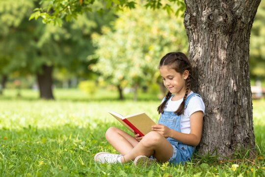Young girl reading book against tree in park setting