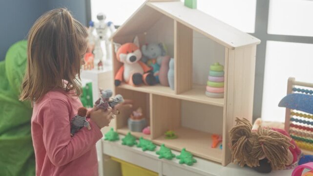 Girl holds doll beside a wooden dollhouse and lines up toy frogs on a shelf in a kids playroom building; playful curiosity.