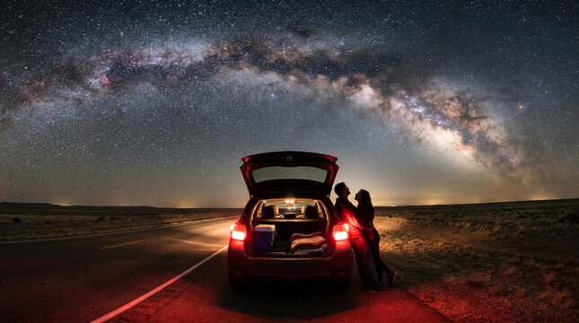 Couple stargazing beside open car boot on empty desert highway under breathtaking Milky Way galaxy arch at night, road trip adventure and romance concept