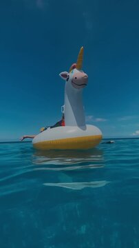 Mother son inflatable unicorn float in crystal ocean under bright blue sky, mother steadying float while child laughs, gentle waves, sunlit reflections, vibrant summer colors, vertical video
