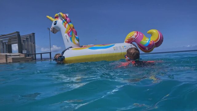 White boy reaching inflatable unicorn while splashing in shallow ocean near dock, wearing goggles and red rashguard, hauling colorful float with playful effort and summer