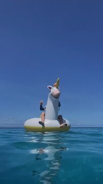 Boy on tipped inflatable unicorn watches float drift across calm blue ocean, gentle ripples, quiet sunlight, distant horizon, solitary drift with playful toy slowly tilting and bobbing, Vertical video