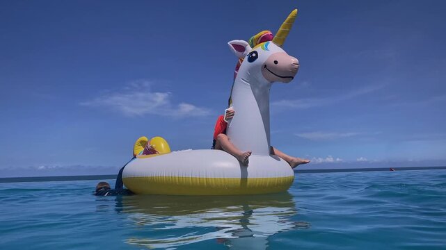Child perched on inflatable unicorn, small silhouette against vast blue sky, toes dangling, lifevest visible, playful solo moment and confident balance on calm sea