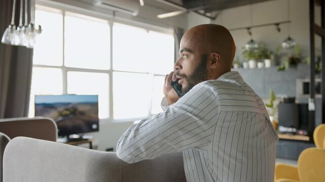 Mixed-race businessman in a modern, sunlit office communicates actively on his mobile phone, leaning casually against a light gray couch, demonstrating professional engagement during daytime hours