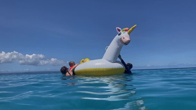 Two kids on unicorn float in wide turquoise ocean, distant horizon and clear sky, gentle swell, carefree exploration, reflective water and bright summer atmosphere