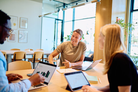 Diverse business professionals having a meeting, analyzing data with laptops and tablets, sharing ideas in a modern collaborative workspace
