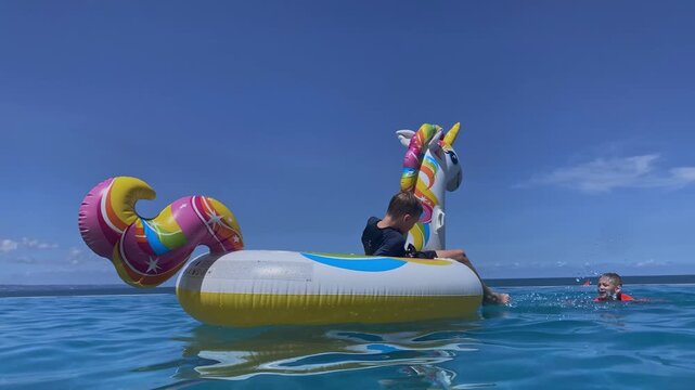 White boy reaching inflatable unicorn while splashing in shallow ocean near dock, wearing goggles and red rashguard, hauling colorful float with playful effort and summer
