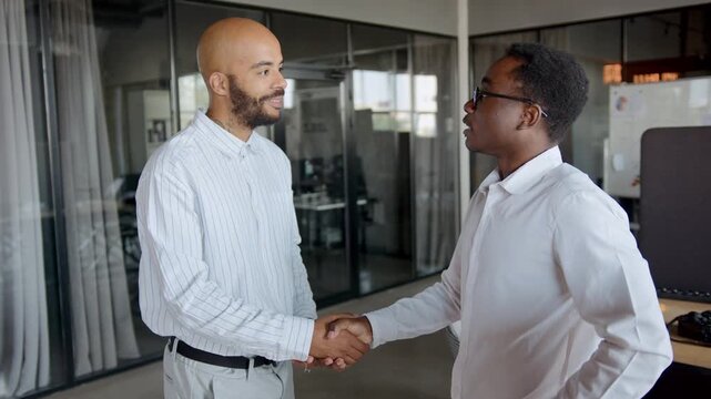 Two men in white shirts shake hands in an office showing teamwork and respect in a workplace setting while discussing their projects