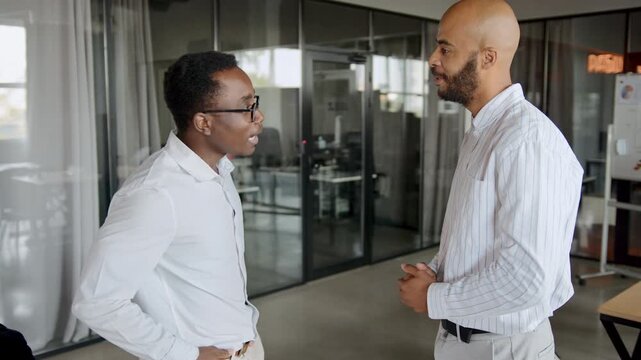 Professional Black men in white shirts greet each other with a handshake in a modern office discussing current business matters and workplace topics during a productive workday