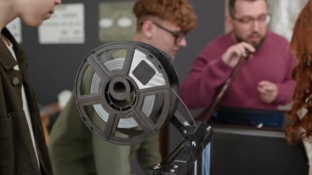 Close up shot of spool of gray filament being fed into 3D printer while group of students and their teacher observing another device in classroom