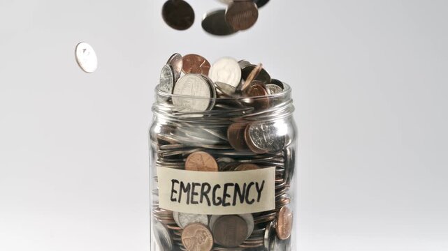 Close-up shot of coins falling into an 'Emergency Fund' coin jar in slow motion.