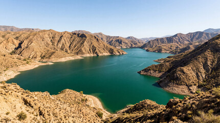 Fototapeta premium Deep Emerald Reservoir Basin Bordered by Sun Baked Clay Hills Under Dramatic Clouds Today