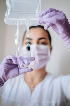 Masked nurse carefully holding syringe near IV bag while preparing intravenous treatment in a bright medical clinic.